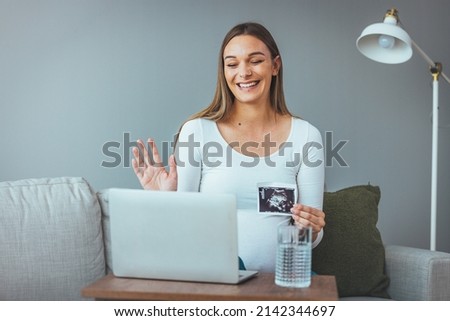 A happy sweet pregnant woman sits on a sofa in the evening and shows an ultrasound picture of her unborn child. He talks on his laptop because he doesn't want contact because of the covid19 virus