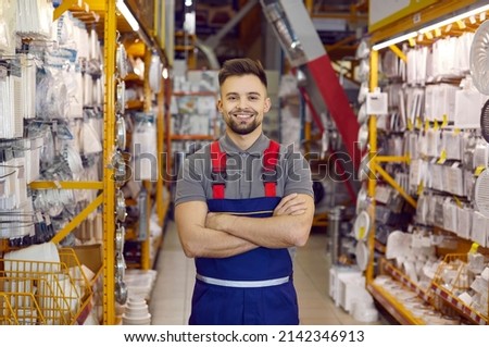 Caucasian consultant in uniform at hardware store. Male standing next to shelves with tools at DIY store. Handsome bearded guy is ready to help customers to choose tools for different home repairs Royalty-Free Stock Photo #2142346913