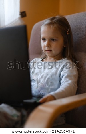 Child watching cartoons on laptop. Serious kid with technology at home in armchair spending time on-line. Education and entertainment for toddlers. Girl in cozy atmospheric room
