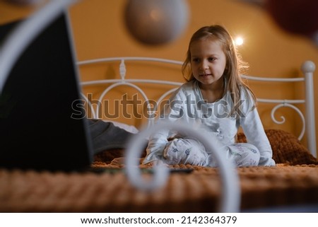 Child watching cartoons on laptop. Serious kid with technology at home on bed spending time on-line. Education and entertainment for toddlers. Girl in cozy atmospheric room over the window