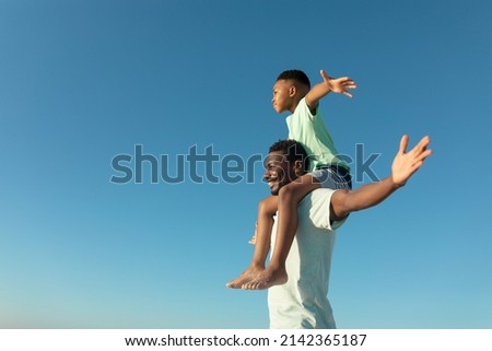 Happy african american boy on father's shoulders by copy space against clear blue sky. unaltered, family, lifestyle, togetherness, enjoyment and holiday concept. Royalty-Free Stock Photo #2142365187
