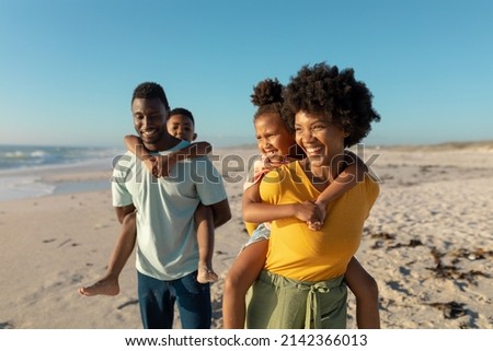 Happy african american parents giving piggyback to children while walking at beach on sunny day. unaltered, family, lifestyle, togetherness, enjoyment and holiday concept. Royalty-Free Stock Photo #2142366013