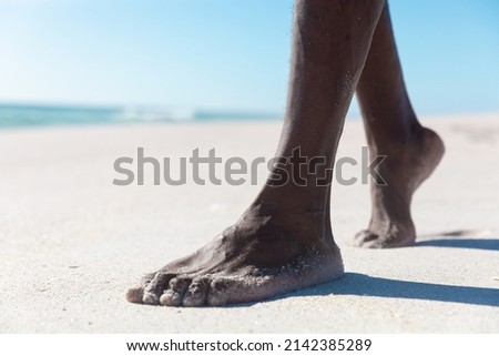 Low section of african american senior man walking barefoot on sand at beach during sunny day. unaltered, active lifestyle, enjoyment and holiday concept. Royalty-Free Stock Photo #2142385289