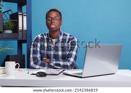 Funny and goofy office working person making amusing playful face while sticking out tongue. Childish and dumb young adult man with dullard face expression while in company workspace. Royalty-Free Stock Photo #2142386819
