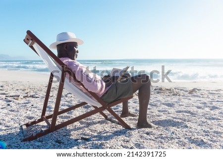 Side view of african american senior man reading book sitting on folding chair at beach. unaltered, hobbies, active lifestyle, enjoyment and holiday concept. Royalty-Free Stock Photo #2142391725