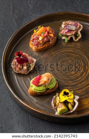 Round brown plate with 5 tapas with beef roast beef, tartare, salmon, chicken and avocado. A dish of haute cuisine. Dark gray textured background. Close up. Royalty-Free Stock Photo #2142395743