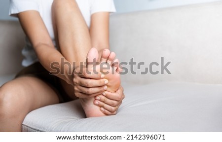 Foot pain, Asian woman sitting on sofa feeling pain in her foot at home, female suffering from feet ache use hand massage relax muscle from soles in home interior, Healthcare and podiatry medical Royalty-Free Stock Photo #2142396071