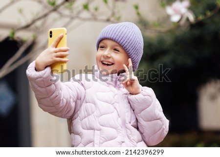 Cute little caucasian girl with blonde hair in trendy casual clothes using mobile phone outdoor at spring. Video chat and social distancing during coronavirus covid-19 pandemic lockdown