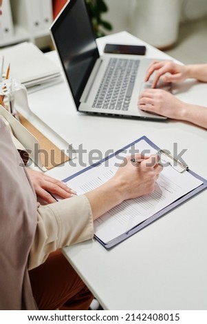 Hand of young female patient filling in medical document in hospital while sitting by desk in front of clinician consulting online patients