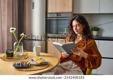 Young relaxed woman in ochre brown blouse and white linen pants reading book while sitting by kitchen table after breakfast Royalty-Free Stock Photo #2142408183