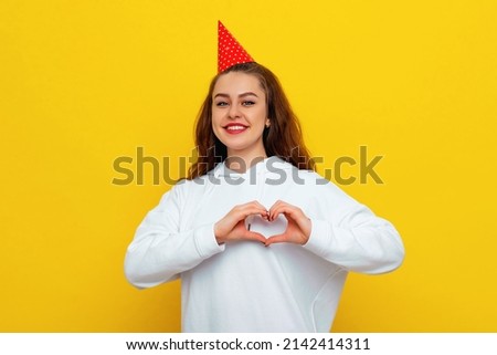 Lovely smiling adorable woman in a red celebratory cap showing heart sign and smiling, express sympathy, care or gratitude, standing yellow background