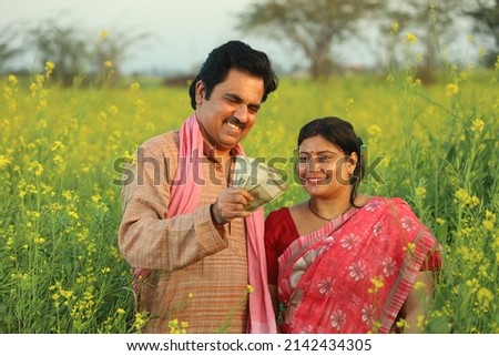 Young happy Indian farmer couple standing in mustard field in a day time enjoying the agricultural profits. They are happy to get benefitted by the mustard flourishing crops and production.  Royalty-Free Stock Photo #2142434305