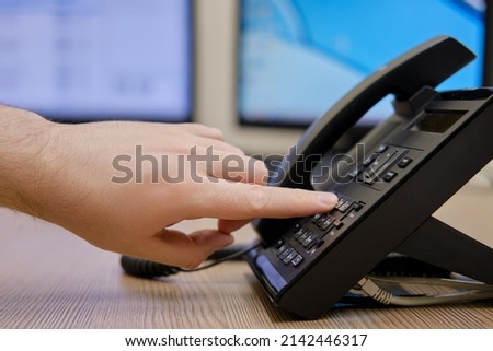 A man hand dials a number on the phone at the monitors of office computers. Finger presses on the buttons of a office landline phone Royalty-Free Stock Photo #2142446317