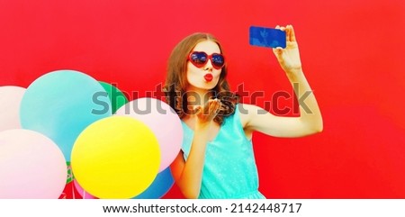 Portrait of woman blowing her red lips sending sweet air kiss taking selfie by smartphone with colorful balloons on red background