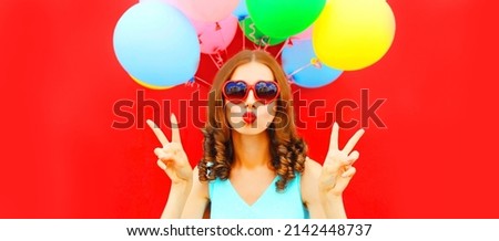 Portrait of beautiful woman blowing her red lips sending air kiss with colorful balloons on red background