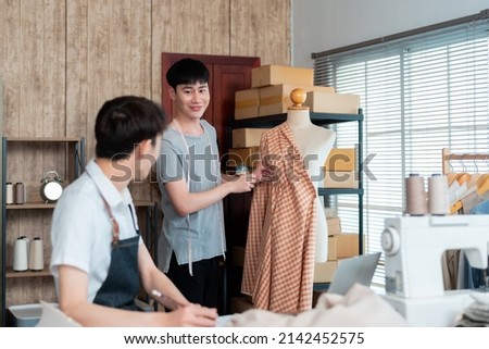 Young LGBTQ Asian Men Small Business PartnerSit and design the dress with a smiling face, there is a background assistant who is fixing the dress.Professional Design clothes to sell online,