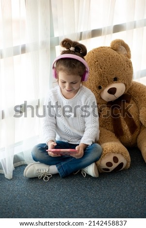 Cute little girl holding tablet computer in her hands. She is in headphones. Advertising new gadget, educational app. She is near the window. Games. Children use technology.