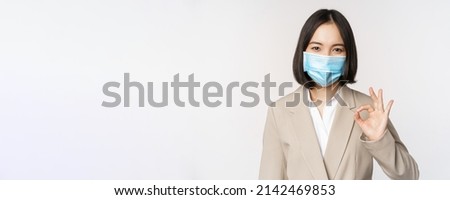 Coronavirus and workplace concept. Image of asian saleswoman, company worker in medical mask showing okay sign, smiling pleased, white background