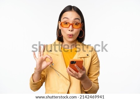 Amazed asian girl in sunglasses, showing okay, ok sign, holding smartphone, looking impressed, recommending smth, standing over white background