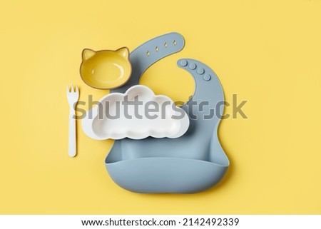 Baby bibs and plate in the shape of a cloud on yellow background. Serving baby. Concept of kids menu, nutrition and feeding  Royalty-Free Stock Photo #2142492339