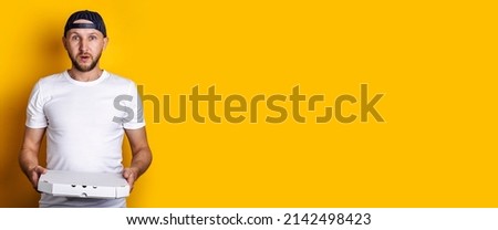 Surprised young man in a baseball cap holding a packaged pizza on a yellow background. Banner.