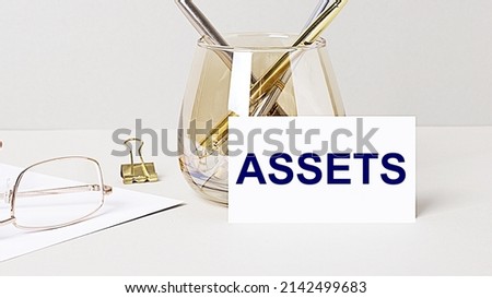 Work desk with gold glasses, pen, white card with the text ASSETS. Business concept. Home Office. Workplace close-up.