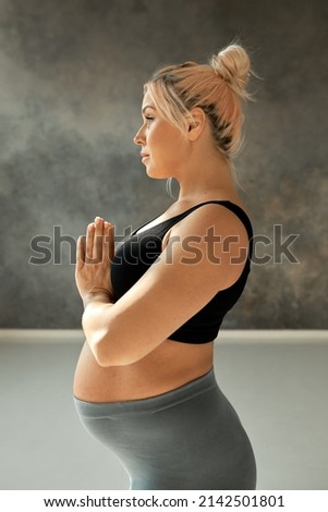 Profile close-up portrait of young pregnant female in sportswear keeping hands in praying position, looking straight in front of her, isolated on grey wall of yoga studio. Fitness and pregnancy