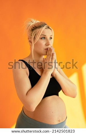 Vertical image of beautiful blonde Caucasian pregnant woman praying with hands together about her labor, her child, looking worried standing against orange sun-drenched studio wall