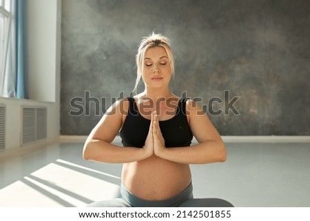 Horizontal picture of pregnant blonde woman meditating, relaxing sitting on floor with hands in namaste gesture, breathing deeply, keeping eyes closed, reaching zen and harmony, getting rid of stress
