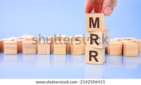 hand holding wood cube block with MRR text. the inscription on the cubes is reflected from the surface. blue background with copy space. MRR - short for Monthly Recurring Revenue
