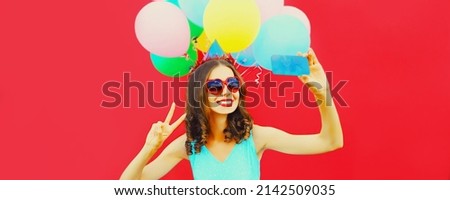 Portrait of happy cheerful smiling woman in birthday hat taking selfie by smartphone with colorful balloons on red background