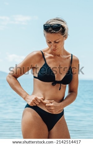 Dissatisfied frowning middle age woman in bikini pinching belly fat while spending day on the beach.