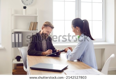 Happy client signing some paper documents. Smiling bearded man in suit sitting at office desk with female bank manager, business assistant, or insurance agency worker and putting signature on contract