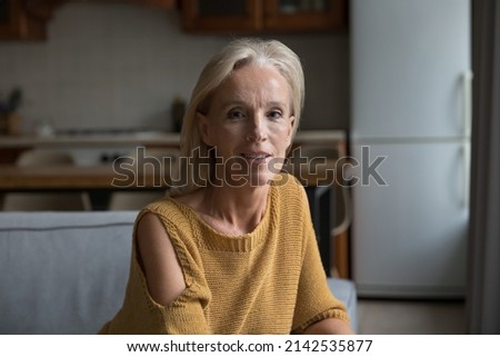 Positive mature lady home head shot portrait. Blonde middle aged pensioner, homeowner, retired woman in casual sitting on couch in living room, looking at camera, smiling