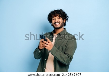 Happy handsome amazed stylish indian or arabian guy, using smartphone, chatting online, texting message, browsing internet, social media, looks at camera, stands on isolated blue background, smiling Royalty-Free Stock Photo #2142569607