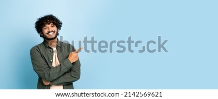 Panoramic photo of arabian or indian guy in casual wear, looks at camera and points finger to the side at empty space for your presentation, stands on isolated blue background, smiling