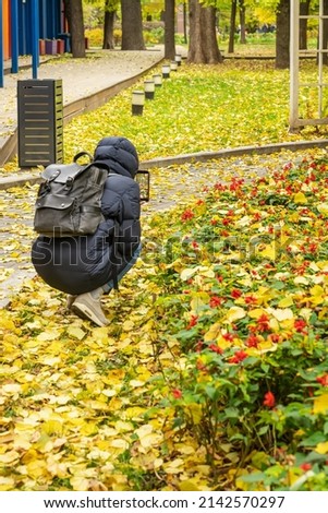 Fall season in city park, leaf fall, young guy taking photo of colorful landscape with fallen leaves and last flowers. Modern technology and autumn nature