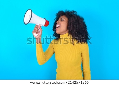 Young woman with afro hairstyle wearing yellow turtleneck over blue background Through Megaphone with Available Copy Space