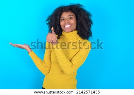 Young woman with afro hairstyle wearing yellow turtleneck over blue background Showing palm hand and doing ok gesture with thumbs up, smiling happy and cheerful.