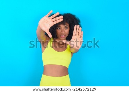 Portrait of smiling young woman with afro hairstyle in sportswear against blue wall looking at camera and gesturing finger frame. Creativity and photography concept.