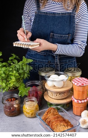 Young white woman dressed in denim apron in the right part writing the weekly list of prepared homemade food. Elegant batch cooking scene. Royalty-Free Stock Photo #2142574407