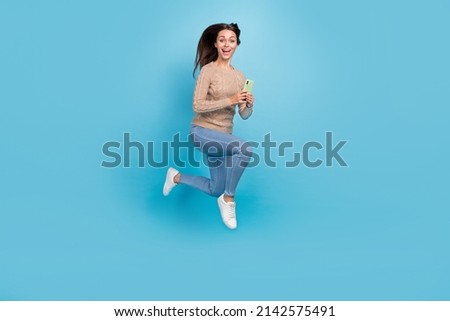 Photo of excited lady jump hold phone read post run wear beige sweater jeans sneakers isolated blue color background