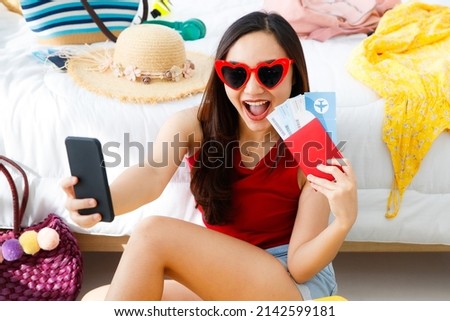 Young happy and excite Asian female traveler getting ready for a summer holiday trip vacation with her bright yellow suitcase wearing heart shape sunglasses taking selfie photo with her passport.