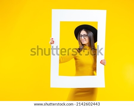 Beautiful woman holds a frame and smiles - isolated over yellow. ?heerful girl in the picture. Woman looks into the camera and smiles. Girl poses with a white frame. Lady in a hat and a yellow dress.