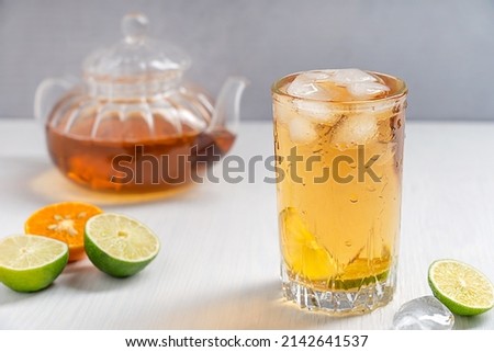 Homemade organic fresh iced tea in tall drinking glass with ice cubes and slices of sour lime and tangerine citrus fruit served on white wooden background with teapot in summer at kitchen for lunch