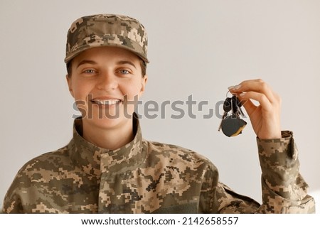 Portrait of smiling positive optimistic military woman wearing camouflage uniform and cap, standing looking at camera with happy smile and holding keys from a new car or apartment. Royalty-Free Stock Photo #2142658557