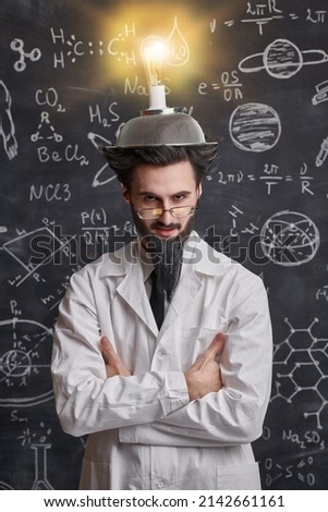 A smart eccentric scientist stands at the blackboard with scientific formulas and diagrams with a strange invention on his head. Science and new inventions. Royalty-Free Stock Photo #2142661161