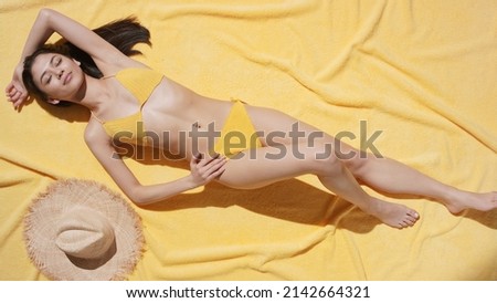Top view shot of young gorgeous slim Asian woman with long brown hair in yellow crochet bikini lying on yellow beach blanket near a straw hat enjoys the sun | UV protection and body care commercial