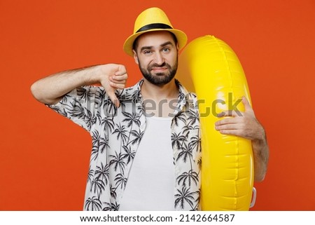 Young disappointed tourist man in beach shirt hat hold inflatable ring showing thumb down dislike gesture isolated on plain orange background studio portrait. Summer vacation sea rest sun tan concept