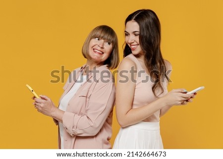Side view two fun young daughter mother together couple women in casual clothes hold mobile cell phone stand back to back look to each other isolated on plain yellow background studio Family concept Royalty-Free Stock Photo #2142664673